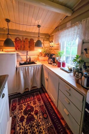 A kitchen with a wooden ceiling, a sink, cabinets, a patterned rug, and a window with lace curtains. - Cozy timber cabin from 1966 with sauna (Kvenvær)