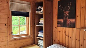 A wooden cabin room with a bed, a window with curtains, a shelf with folded towels, and a wall-mounted deer picture.