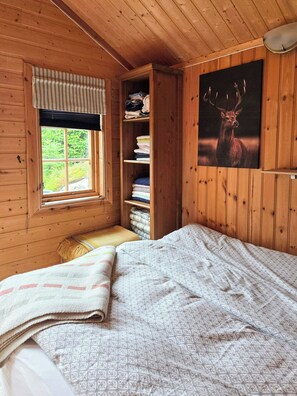 A wooden cabin room with a bed, a window with curtains, a shelf with folded towels, and a wall-mounted deer picture. - Cozy timber cabin from 1966 with sauna (Kvenvær)