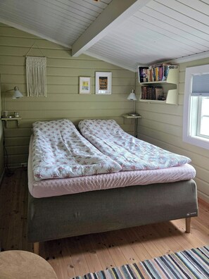A bedroom with a bed, bedside lamps, a macrame wall hanging, framed pictures, a bookshelf, and a window with blinds.