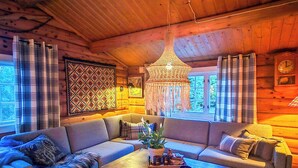 A cozy cabin interior with a wooden ceiling, a large chandelier, a sofa set, a coffee table, and a rug.