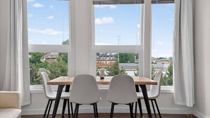Un espace repas comprenant une table et des chaises, de grandes fenêtres avec des rideaux, et une vue sur les bâtiments et la verdure environnante.