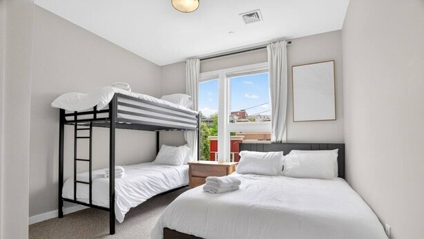 Une chambre avec un lit superposé et un lit simple, tous deux dotés de draps blancs, une fenêtre avec des rideaux et un tableau encadré au mur.