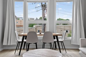 Un área de comedor con mesa y sillas, ventanas amplias con cortinas, y vistas a edificios y líneas eléctricas.
