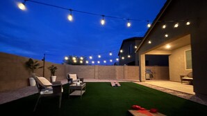 Ein Außenbereich mit Terrasse, Lichterketten, einem Grill und einem grünen Kunstrasen.