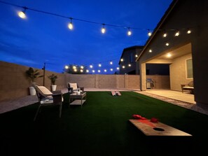 Ein Außenbereich mit Terrasse, Lichterketten, einem Grill und einem grünen Kunstrasen.