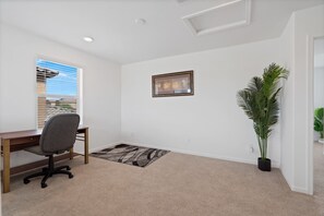 Ein Zimmer mit Schreibtisch, Stuhl, gerahmtem Bild, Topfpflanze und Blick durch ein Fenster.