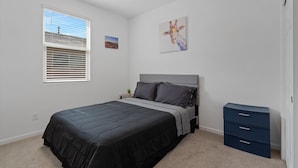 Ein Schlafzimmer mit Bett, Nachttisch, Schminktisch, einem Fenster mit Jalousien und einem Wandbild.