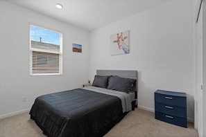 A bedroom with a bed, a nightstand, a dresser, a window with blinds, and a wall with a painting.