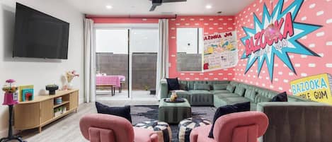 Een woonkamer met een bank, twee roze stoelen, een televisie en een muur met een kleurrijke muurschildering.