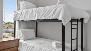 Un lit superposé avec une literie blanche et une commode en bois, dans une pièce dotée d’une fenêtre.