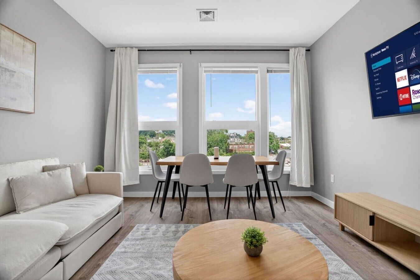 A modern living room with a sofa, a dining table with chairs, a round coffee table with a plant, and a flat-screen TV mounted on the wall.