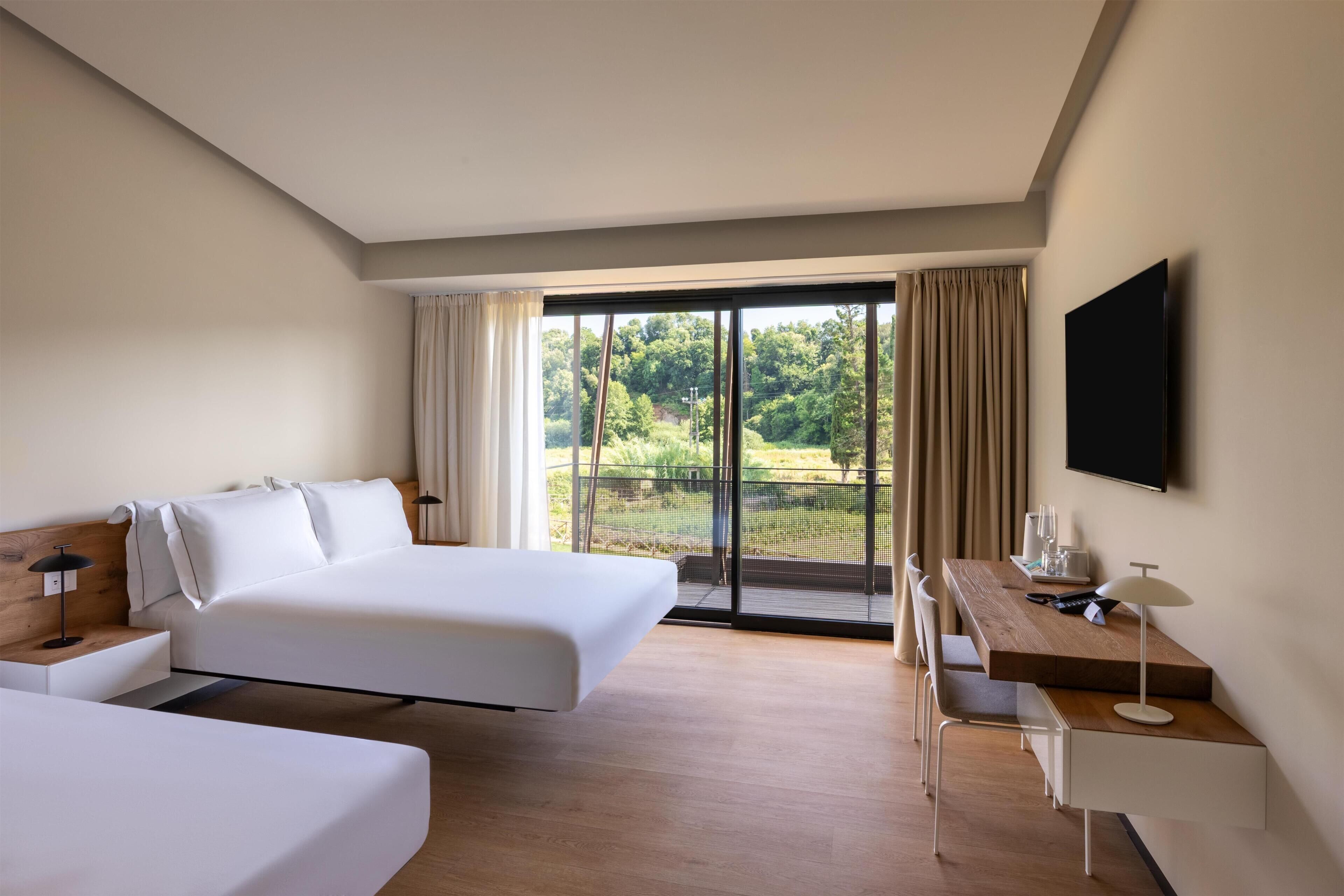 A hotel room with two beds, a desk, a chair, a TV, and a balcony with a view of greenery.