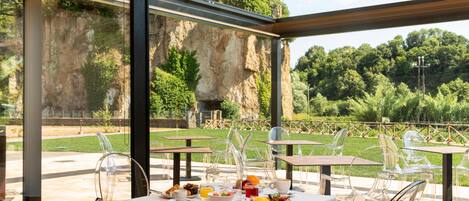 An outdoor dining area with tables and chairs, a view of a rocky cliff and greenery, and a clear sky.
