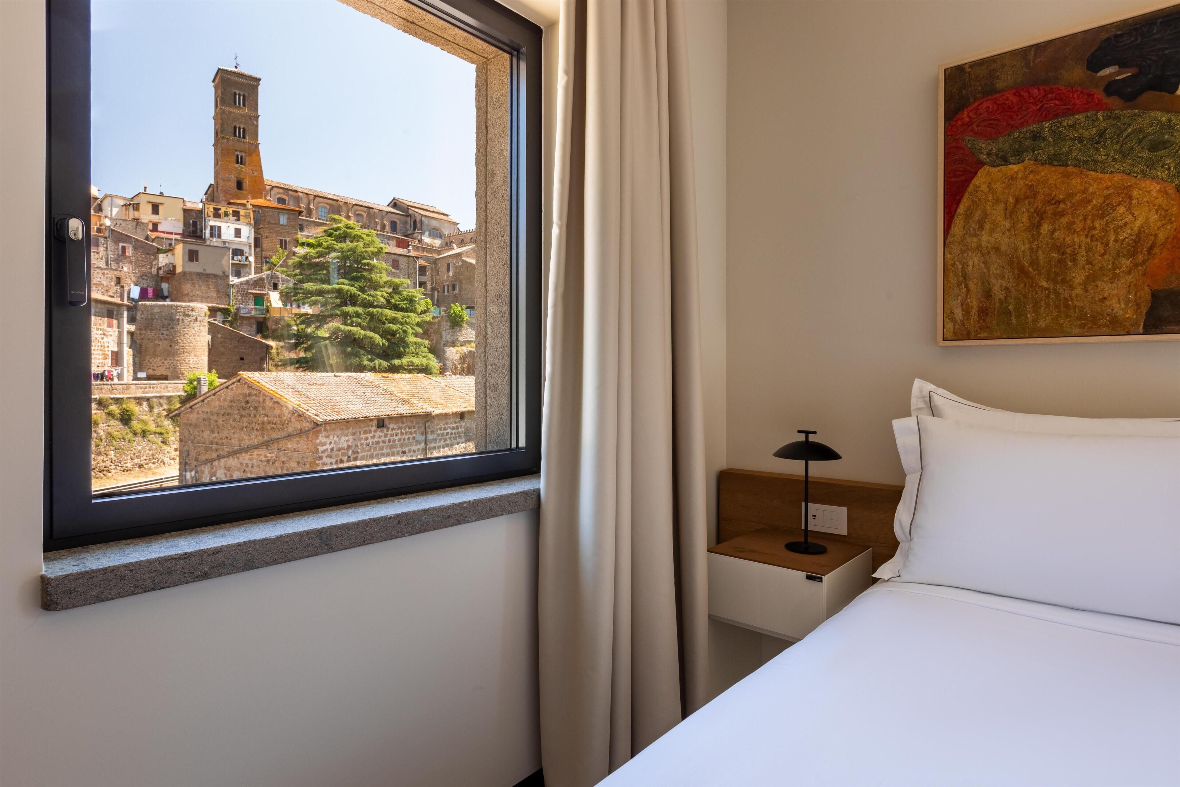 A hotel room with a bed, a nightstand, a lamp, a painting, and a window with a view of historic buildings.