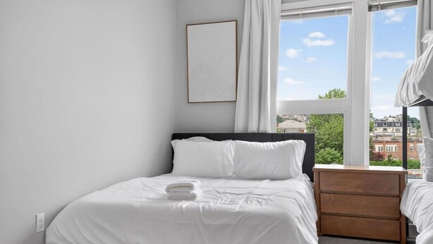 Une chambre d’hôtel avec un lit, une table de chevet, une fenêtre donnant sur des immeubles et un tableau encadré au mur.