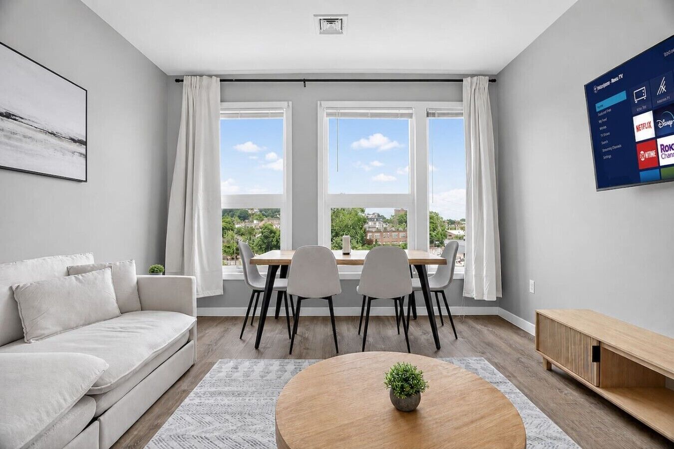 A modern living room with a sofa, a dining table with chairs, a coffee table with a plant, and a TV mounted on the wall.