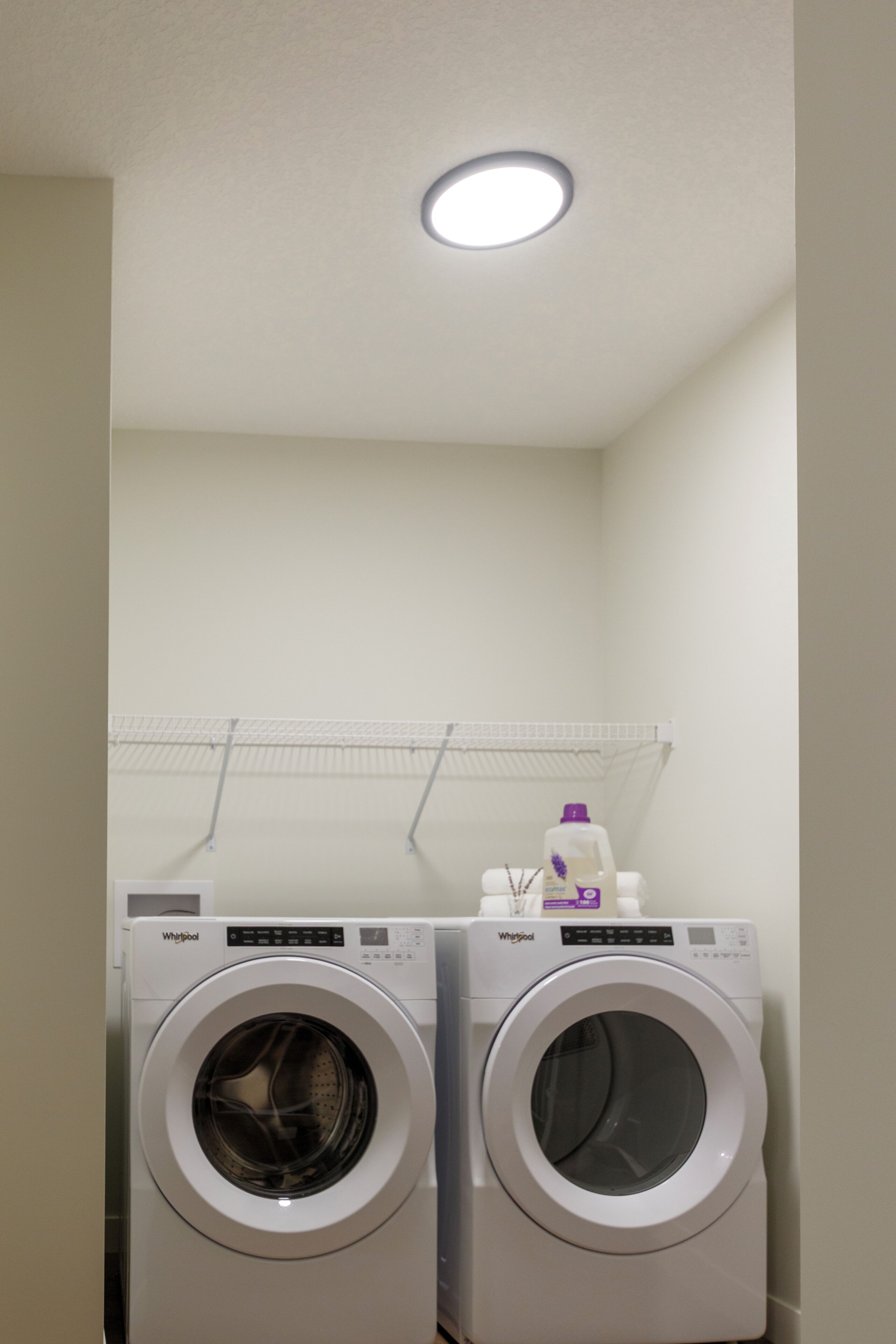A laundry room with two Whirlpool washing machines and a shelf above them.