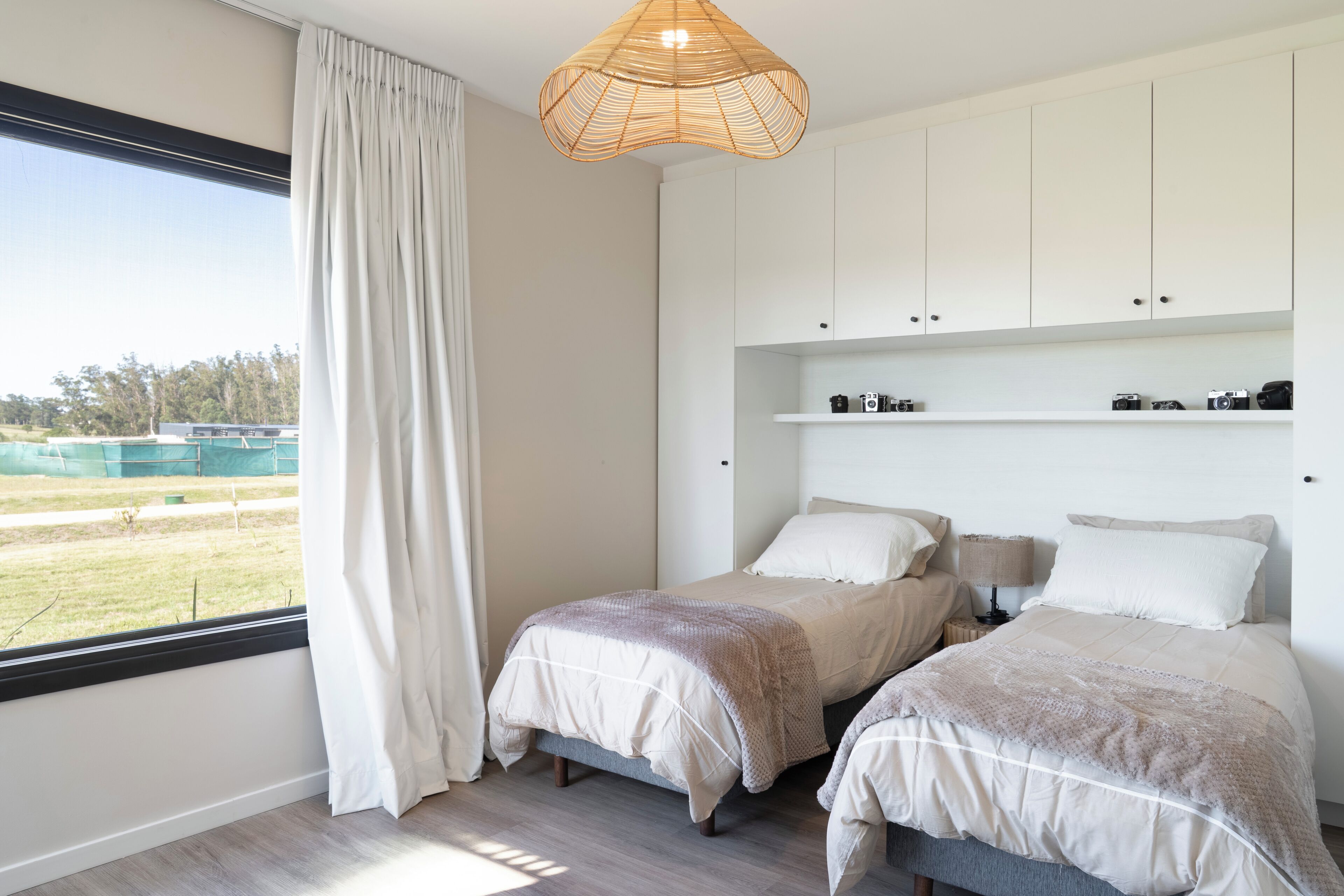 Une chambre avec deux lits, une grande fenêtre avec des rideaux, un luminaire suspendu et une étagère avec des appareils photo.