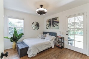 Une chambre à coucher avec un lit, une table de chevet, une plante en pot, un miroir et des œuvres d’art accrochées au mur.