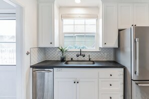 A kitchen with white cabinets, a stainless steel refrigerator, a window with blinds, and a tiled backsplash. - West LA Beachy Bungalow (Los Angeles)