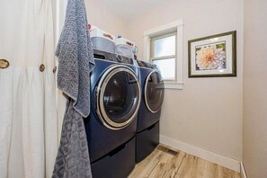 A laundry room with two front-loading washing machines, a white door, a window, and a framed floral artwork on the wall. - Historic Lakefront Farmhouse on Battle Lake - Sleeps 9 (Battle Lake)