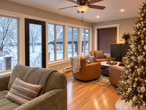 A cozy living room with a fireplace, a Christmas tree, and large windows offering a view of a snowy landscape. - Historic Lakefront Farmhouse on Battle Lake - Sleeps 9 (Battle Lake)