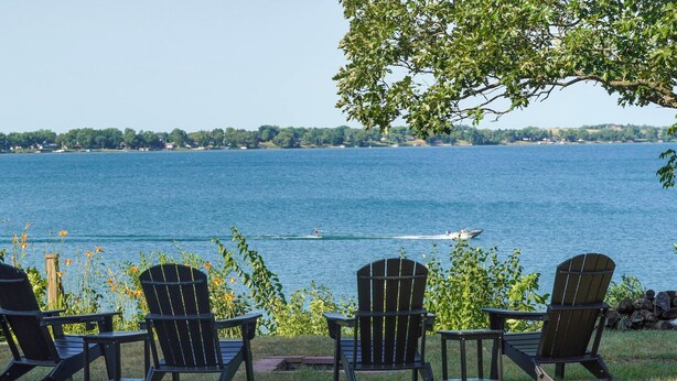 Blick auf einen See mit vier Adirondack-Stühlen, die zum Wasser zeigen, eine Baumreihe und ein Boot in der Ferne.
