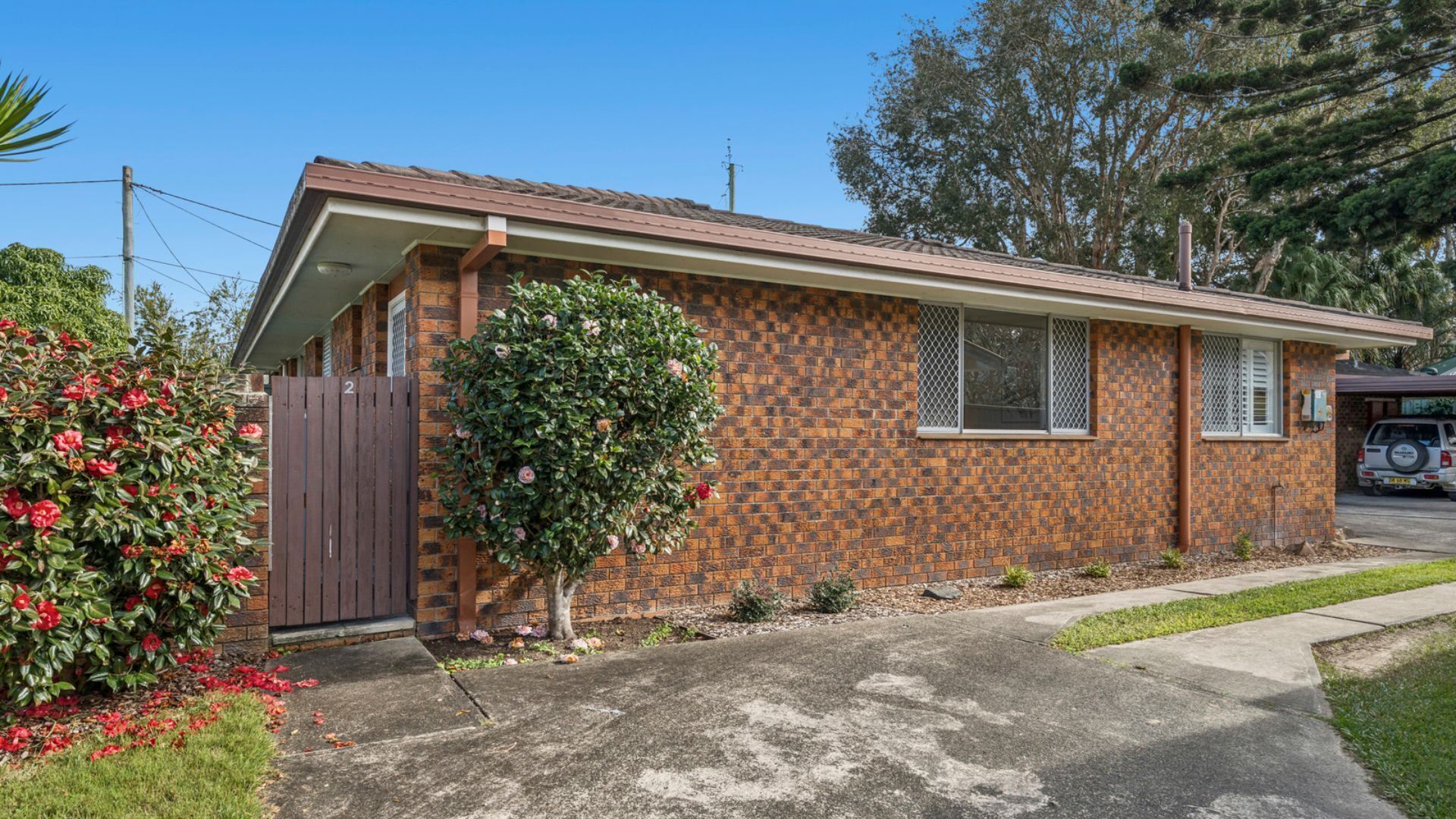 A single-story brick house with a small front yard, a wooden gate, and a bush with red flowers.