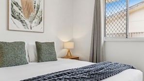 Un lit avec une couette blanche, deux oreillers verts, un plaid bleu foncé en tricot, une lampe de chevet et une estampe botanique encadrée accrochée au mur.
