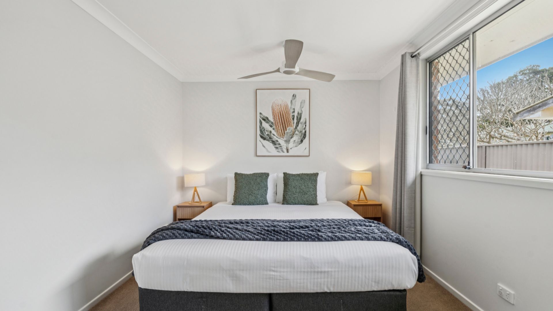 A bedroom with a bed, two bedside lamps, a ceiling fan, a framed picture on the wall, and a window with a view.
