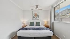 A bedroom with a bed, two bedside lamps, a ceiling fan, a framed picture on the wall, and a window with a view.