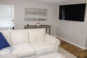 A living room with a white sofa, a small wooden table, a painting, and a television mounted on the wall. - Serendipity Cottage (Prescott)