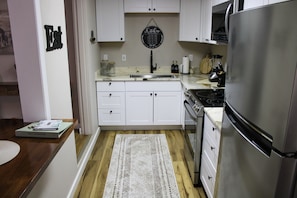 A kitchen with white cabinets, a stainless steel refrigerator, and a black wall clock. - Serendipity Cottage (Prescott)