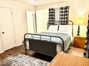 Quarto com estrutura de cama de metal, mesa de cabeceira com abajur, mesa de apoio de madeira, tapete estampado e uma porta.