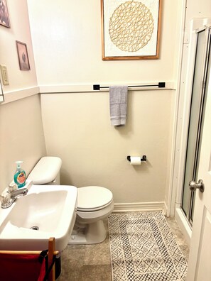 A bathroom with a toilet, sink, and shower. There is a towel hanging on a rod, a picture on the wall, and a patterned rug on the floor.