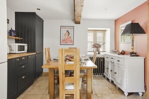 A kitchen with a dining table, chairs, a microwave, cabinets, a radiator, and a window with blinds. - The Tractor Shed: A Quiet Retreat Near Symonds Yat (Ross-on-Wye)