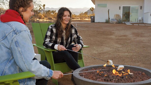 Two people sitting on green chairs outdoors, roasting marshmallows over a fire pit. - Sage Space By Homestead Modern (Yucca Valley)