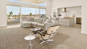 A modern interior with a large L-shaped sofa, a dining area, and a kitchen with a view of the outdoors. - Sage Space By Homestead Modern (Yucca Valley)