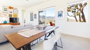 Ein modernes Wohnzimmer mit einer Couch, einem Esstisch und Stühlen, einem Fernseher, Kunstwerken an den Wänden und einem großen Fenster mit Blick auf eine Wüste.
