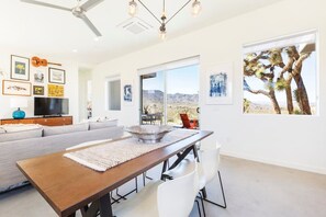 A modern living room with a sofa, dining table, and chairs, a television, artwork on the walls, and a large window with a desert view. - Shelter 1 by Homestead Modern (Joshua Tree)