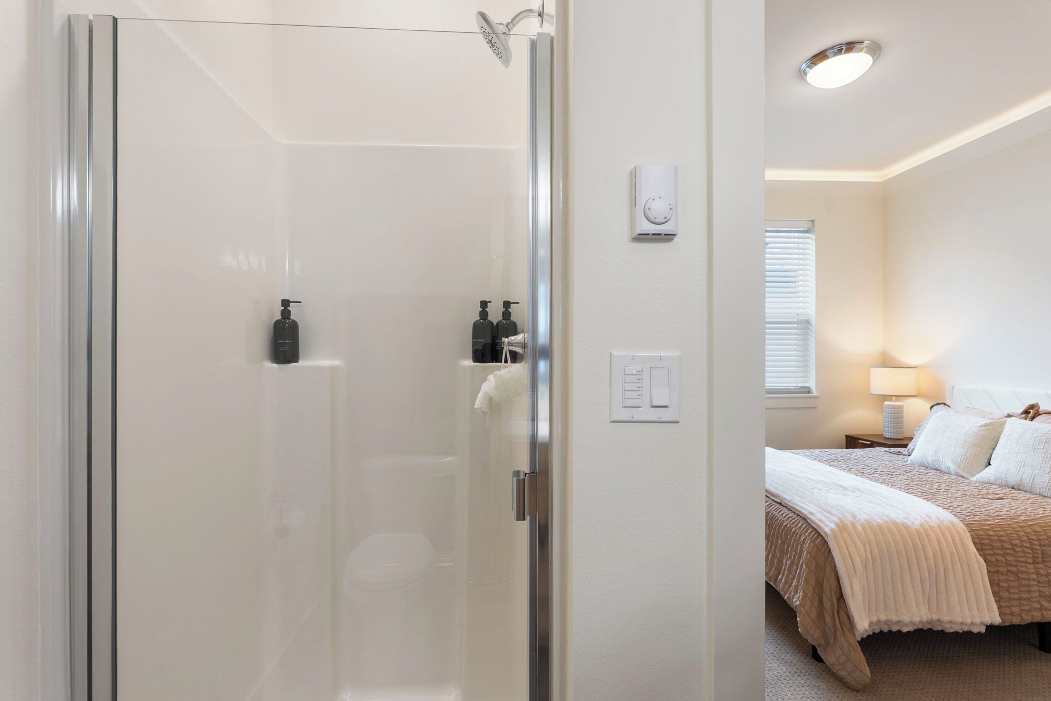 Een kamer met een douche in een glazen cabine, een bed met een beige sprei, een nachtkastje met een lamp en een wandthermostaat.