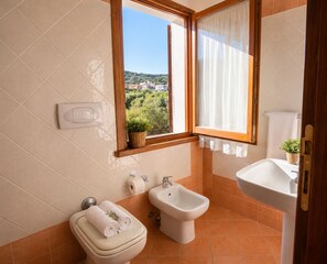 A bathroom with a toilet, bidet, sink, and a window with a view of greenery. - Charming 2-bedroom House in Gallura, North-east Sardinia (Santa Teresa di Gallura)
