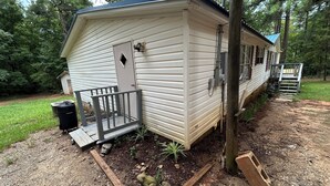 A small, single-story mobile home with a porch, a small shed, and a garden area with a frog statue. - Adorable 3-bedroom  home great for fishing on a private lake in Central Georgia (Macon)