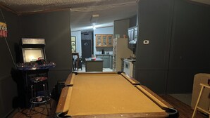A pool table in a room with a dark wall, a small bar area with stools, and a kitchen in the background. - Adorable 3-bedroom  home great for fishing on a private lake in Central Georgia (Macon)