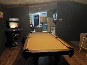 Ein Billardtisch in einem Raum mit dunkler Wand, eine kleine Bar mit Hockern und eine Küche im Hintergrund.