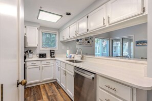A modern kitchen with white cabinetry, a stainless steel dishwasher, and a window with blinds. - Steps to Beach | Palmetto Dunes Pool & Hot Tub (Hilton Head Island)