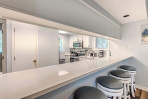 A modern kitchen with a long countertop, bar stools, and a window with blinds. - Steps to Beach | Palmetto Dunes Pool & Hot Tub (Hilton Head Island)