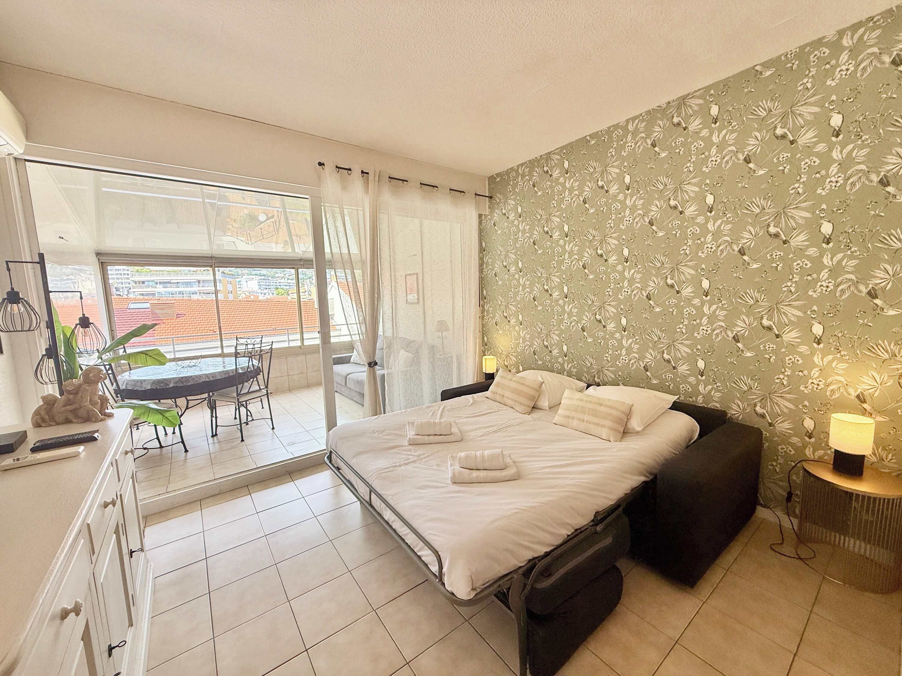 Ein Schlafzimmer mit einem Bett, Nachttischen, einem Fenster mit Vorhängen, einem Balkon und einer Essecke mit Tisch und Stühlen.
