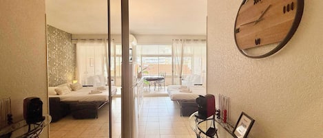 A hotel room with a bed, a sitting area with a table and chairs, a large wall clock, and a view of the outside through a glass door.